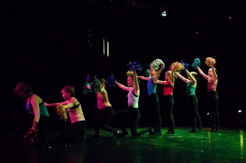 Zespół taneczny skladający się z samych dziewczynek występuje na scenie. Dziewczynki trzymają kolorowe pompony w rękach.