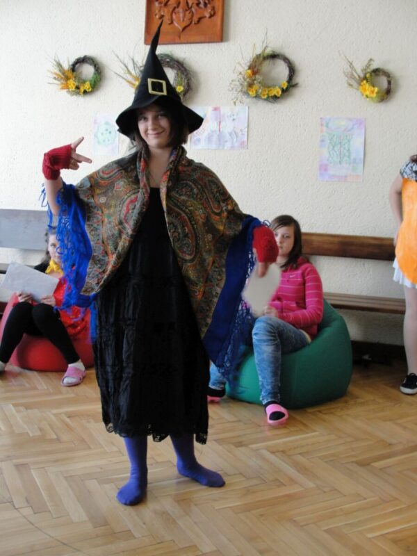 Młoda kobieta przebrana za czarodziejkę, ubrana w czarną pelerynę i długi szpiczasty kapelusz.