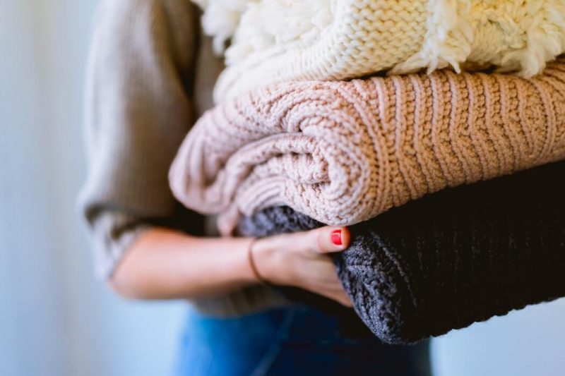 Kobieta trzyma w ręku trzy kolorowe swetry zrobione na drutach