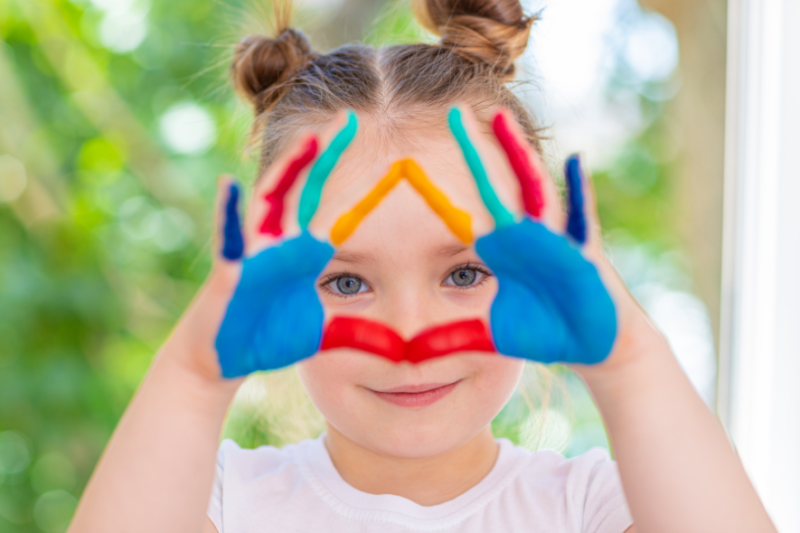 Dziewczynka łączy kciuki i palce wskazujące obu dłoni, które umalowane makolorowymi farbami