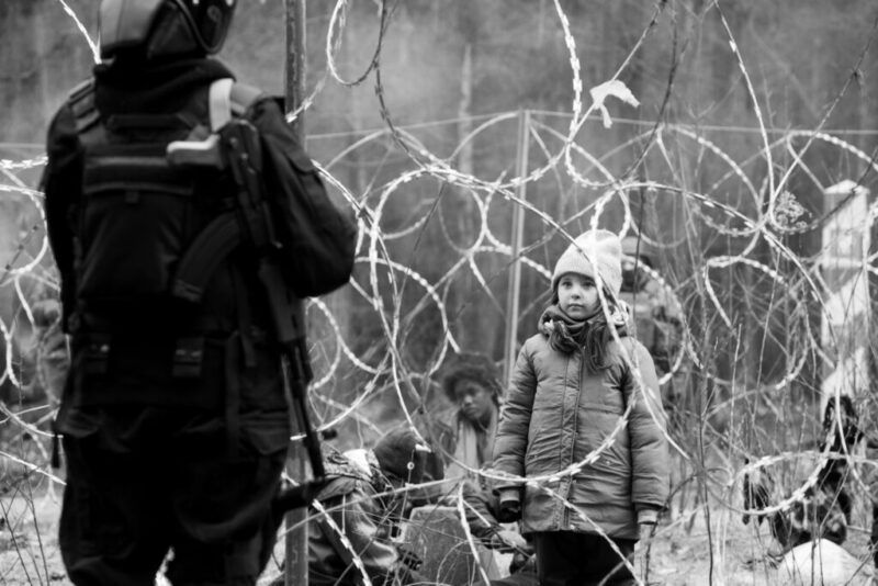 Za kolczastym murem w lesie po jednej stronie stoi dziewczynka, a po stronie polskiej stoi strażnik graniczny