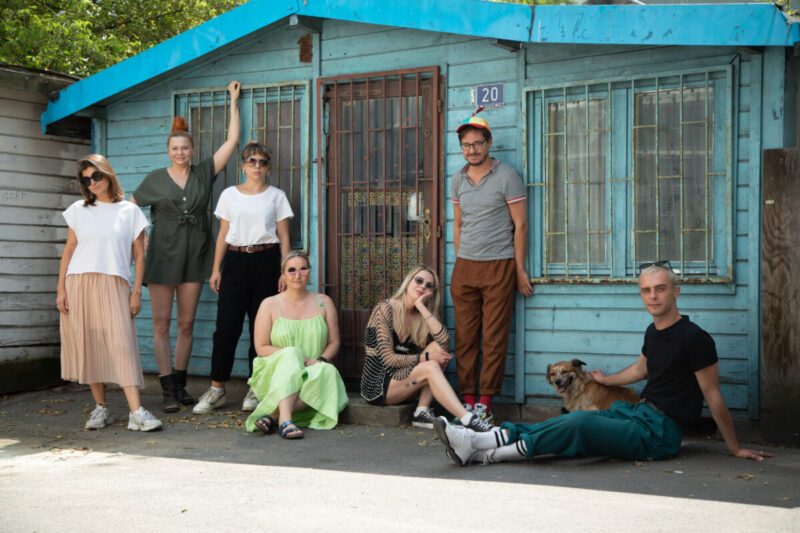 7 osobowa grupa teatralna złożona z osób dorosłych stoi przy zamkniętej, drewnianej, niebieskiej budce - sklepie