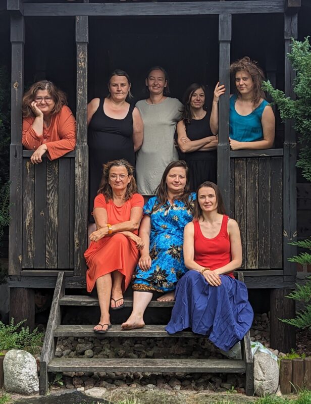 Autor fot. Paweł Misiejuk. 8 - osobowy zespół kobiet, które znajdują się na werandzie drewnianego domu położonego w lesie.