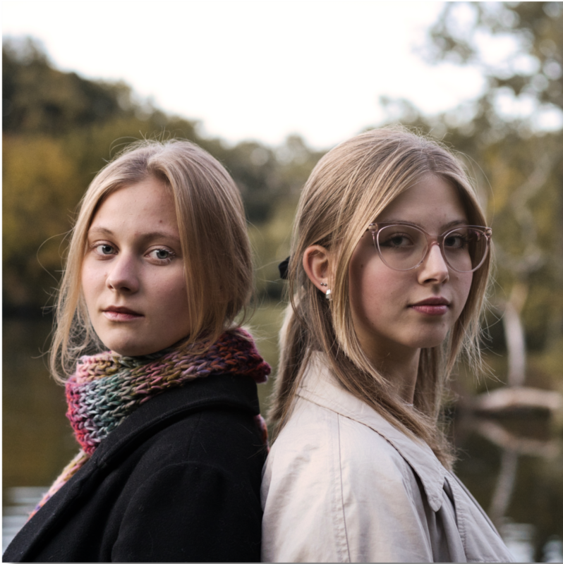 Dwie młode kobiety stoją w parku, opierają się plecami do siebie i patrzą w jednym kierunku, w strone obiektywu