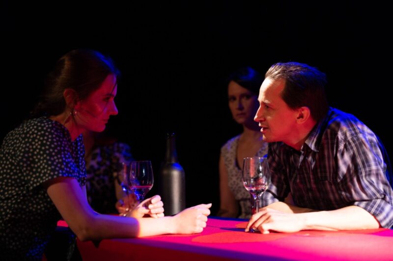 Zdjęcie ze spektaklu THE END przestawiające 3 aktorów, w tym 2 - kobieta i mężczyzna rozmawiają na pierwszy planie, mężczyzna dzierży w dłoni kieliszek i patrzy na kobietę, Zdjęcie jest ciemne.