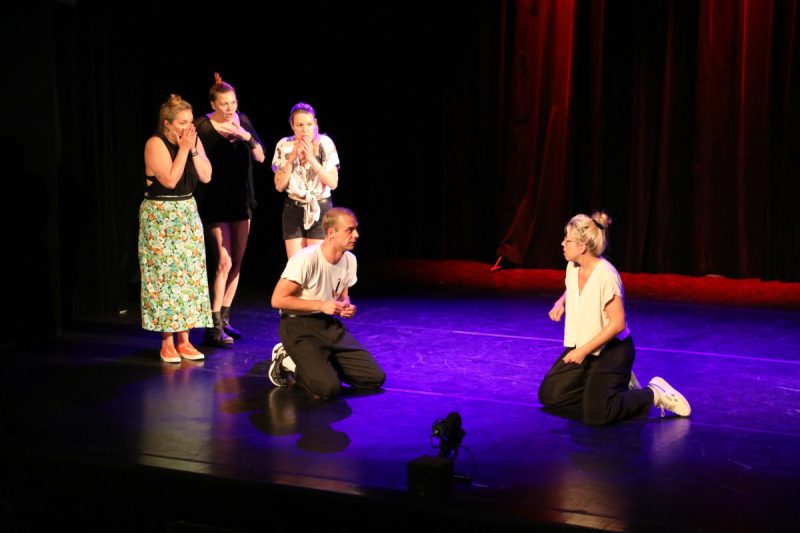na scenie dwie osoby klęczą a, pozostałe 3 osoby stoją z tyłu i każda rękoma zasłania ustami.