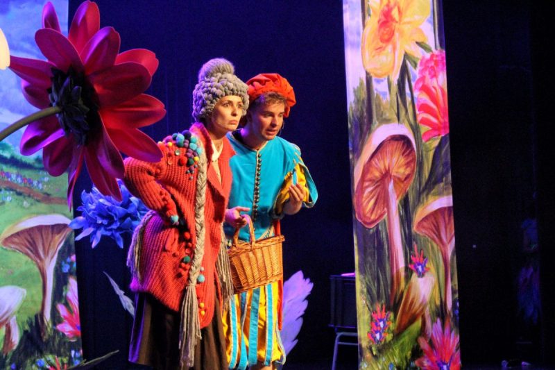 Dwójka aktorów ubranych w kolorowe ubrania z drewnianym koszykiem w ręku stoją na drewnianej scenie między dekoracjami z kwiatów.