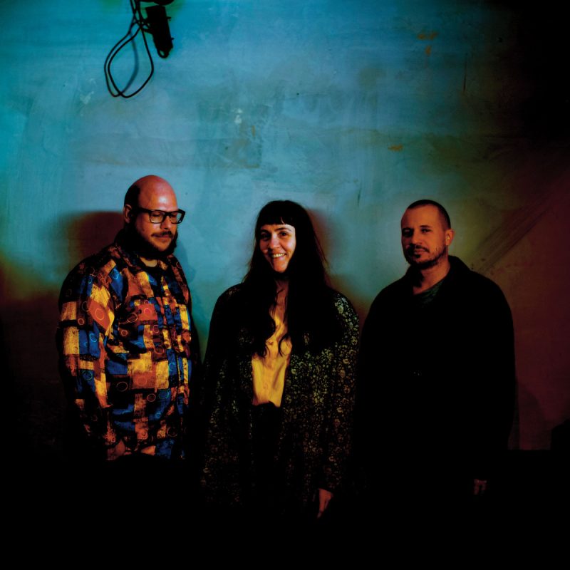 Trzy osoby stojące obok siebie na tle ciemnej, niebiesko-zielonej ściany. Po lewej stronie mężczyzna w kolorowej, wzorzystej koszuli, na środku kobieta z długimi włosami w jasnej bluzce i ciemnym płaszczu, po prawej mężczyzna w ciemnej kurtce. Nad osobami widać przewód i mikrofon wiszące z sufitu.