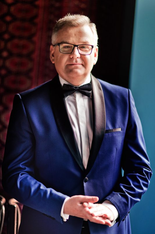 Mężczyzna w białej koszuli i granatowej marynarce, stoi przed obiektywem, trzyma obydwie ręce złożone przed sobą. W tle brązowa ściana.